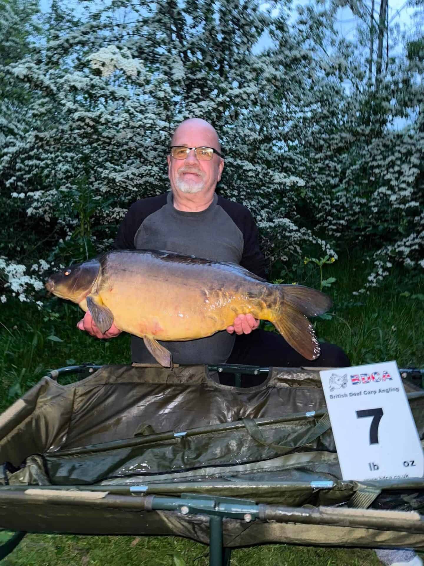 A man wearing glasses holds a large fish, standing outdoors near bushes. A sign in the foreground shows the number 7 and details related to British Steel Carp Angling.