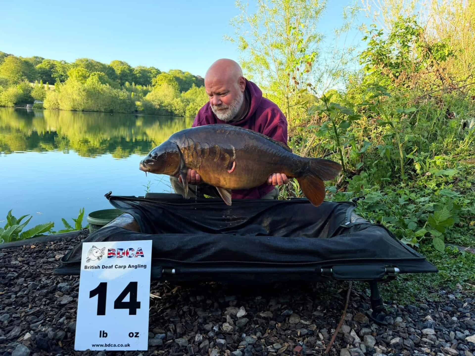 A man holds a large carp by a lakeside, next to a sign reading "14 lb" labelled British Deaf Carp Angling.