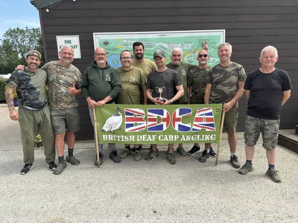 A group of ten men stand together outdoors behind a "British Deaf Carp Angling" banner; one man in the center holds a trophy.