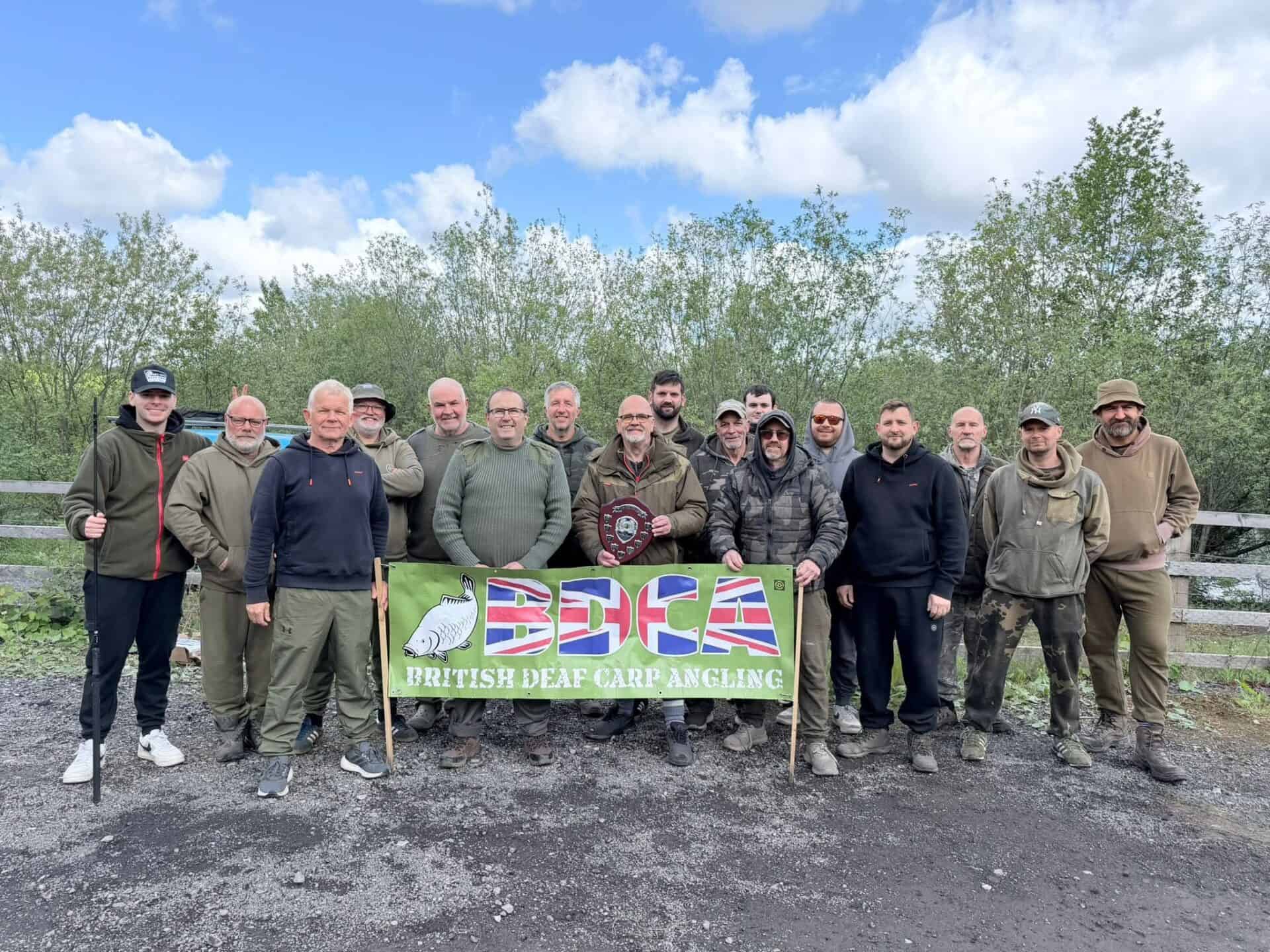 Sixteen people stand outdoors behind a banner reading "British Deaf Carp Angling." One person in the centre holds a plaque. Trees and a wooden fence are in the background.