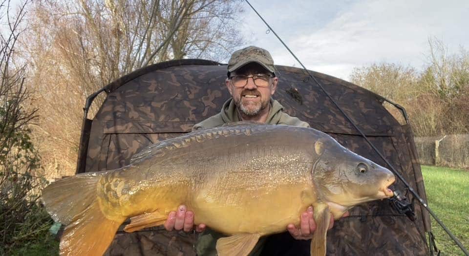 A man wearing glasses and a cap holds a large fish in front of a camouflaged tent, with fishing equipment and trees in the background.