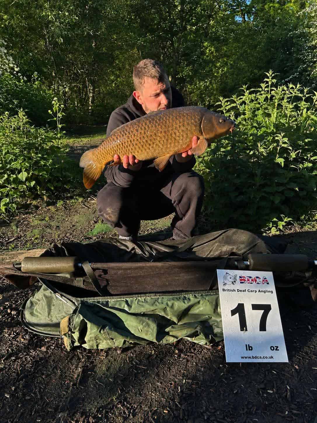 A man kneels outdoors holding a large fish with a sign in front reading "British Dead Carp Angling, 17 lb" next to fishing tackle.