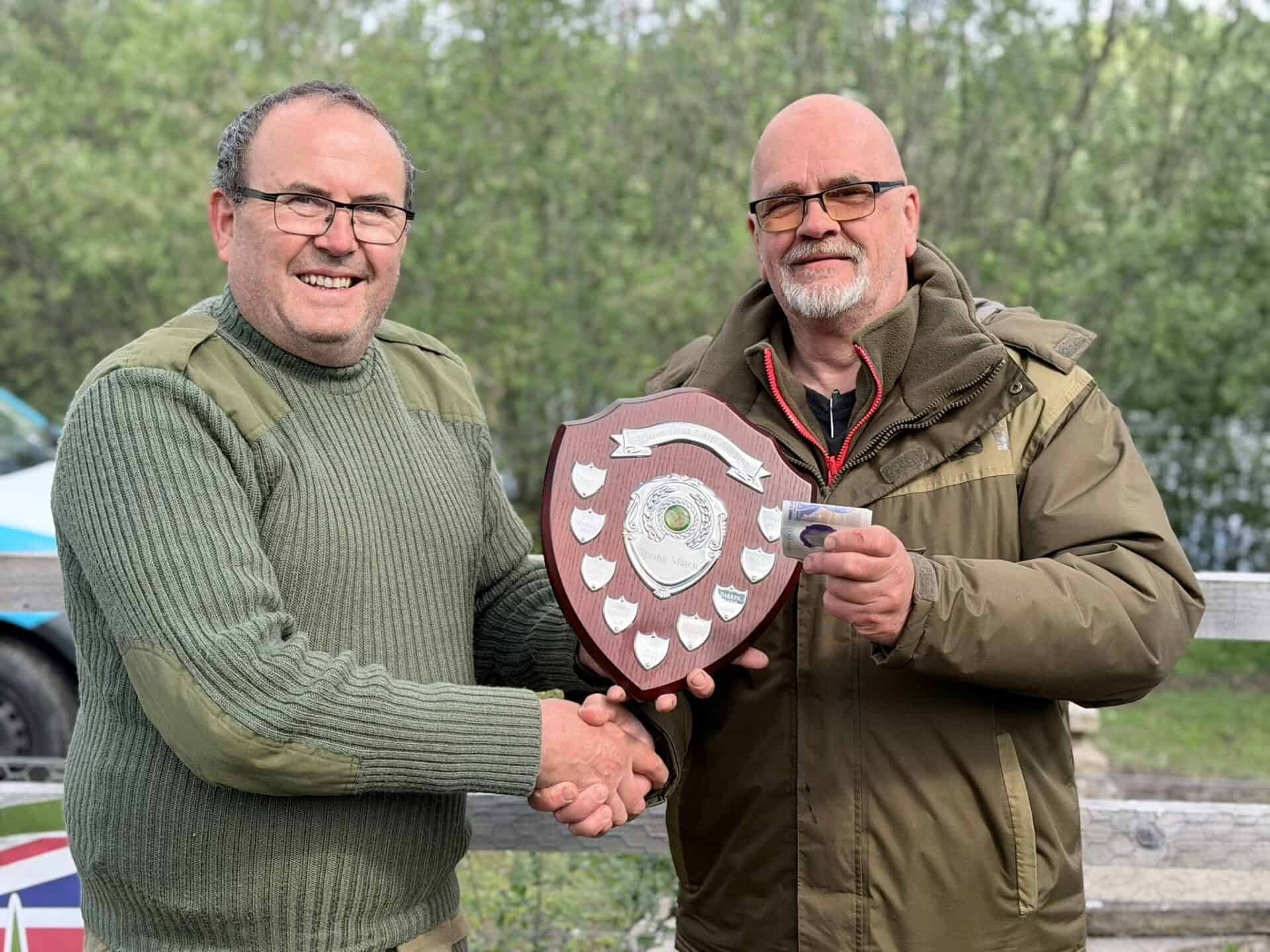 Two men stand outdoors shaking hands. One man holds a shield-shaped award plaque, and the other holds a small rectangular item. Trees and a fence are visible in the background.