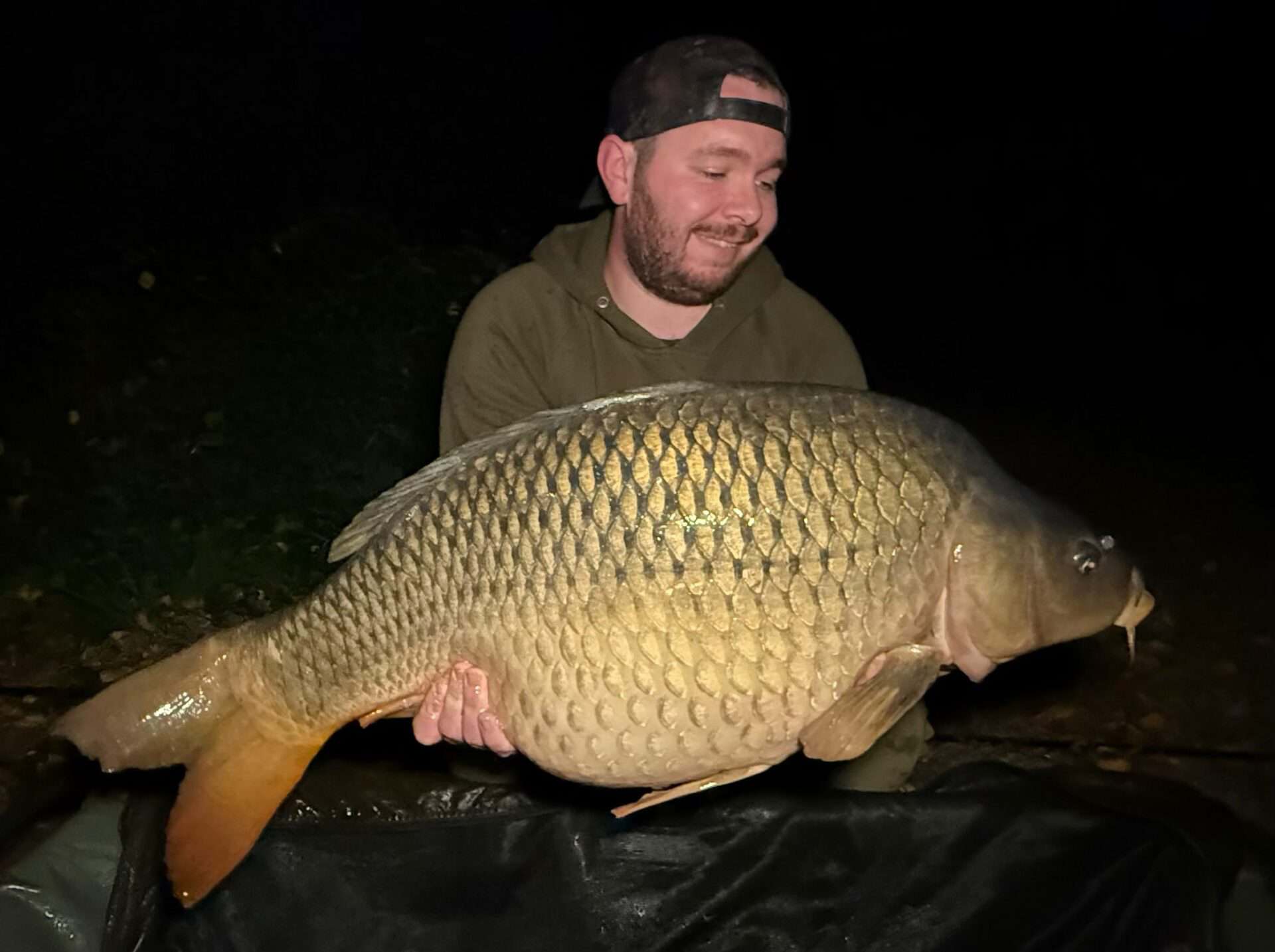 A person wearing a cap and hoodie holds a large fish, likely a carp, at night.