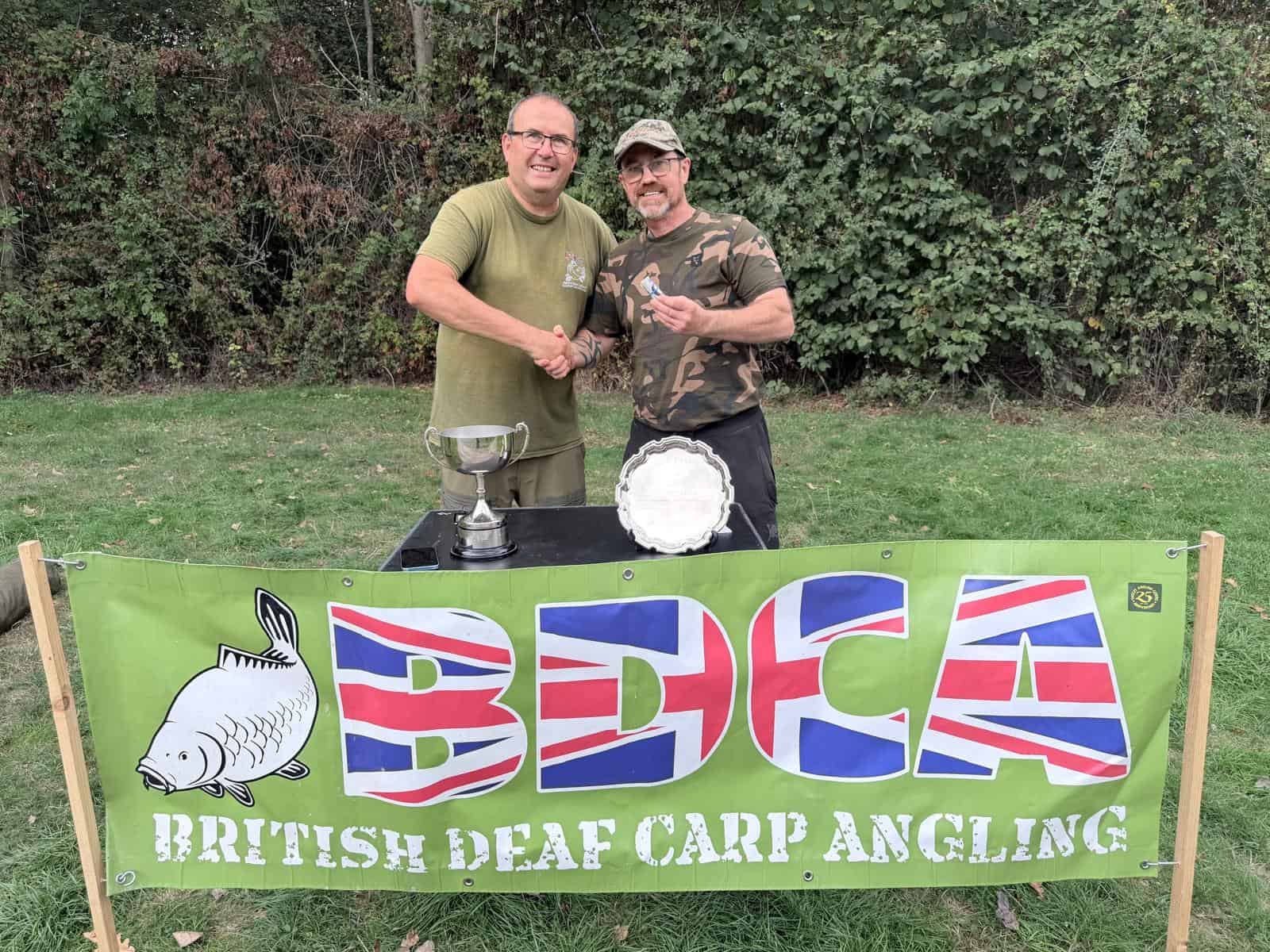 Two men shake hands behind a table with trophies, standing in front of a “British Deaf Carp Angling” banner outdoors.