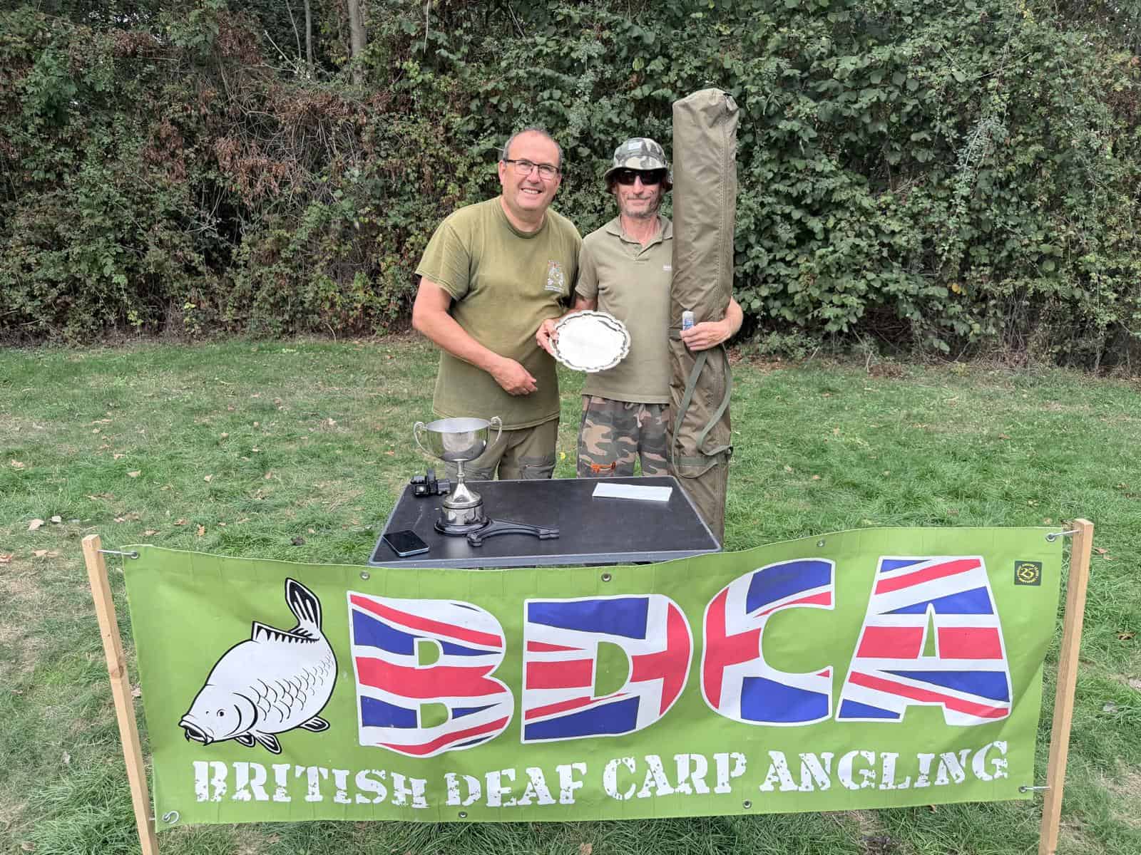 Two people stand behind a table with a trophy and plate, in front of a “British Deaf Carp Angling” banner, with trees in the background.