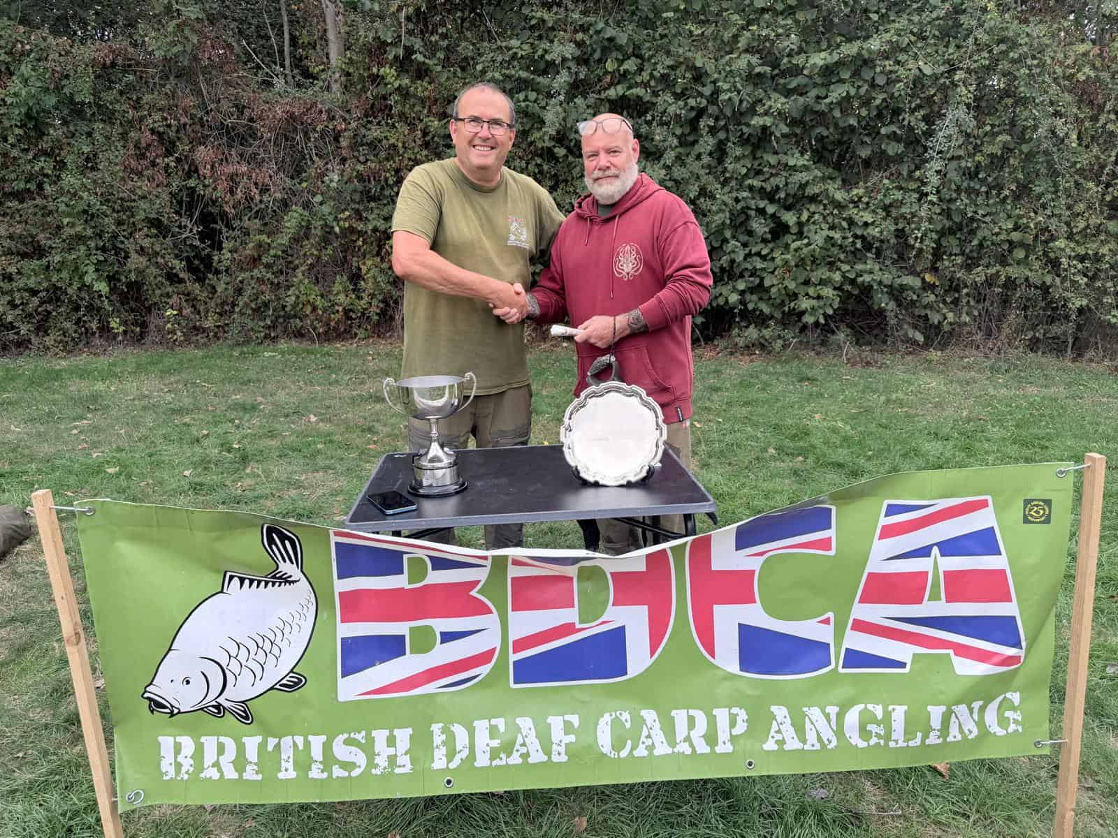 Two men shake hands behind a table with a trophy and a silver platter. A green banner in front reads “British Deaf Carp Angling” with a fish graphic and UK flag letters.