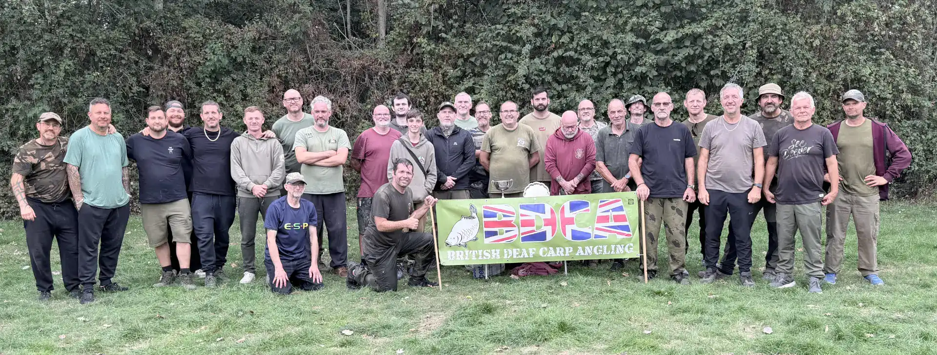 A group of men standing in a park holding a banner that reads “British Deaf Carp Angling”.