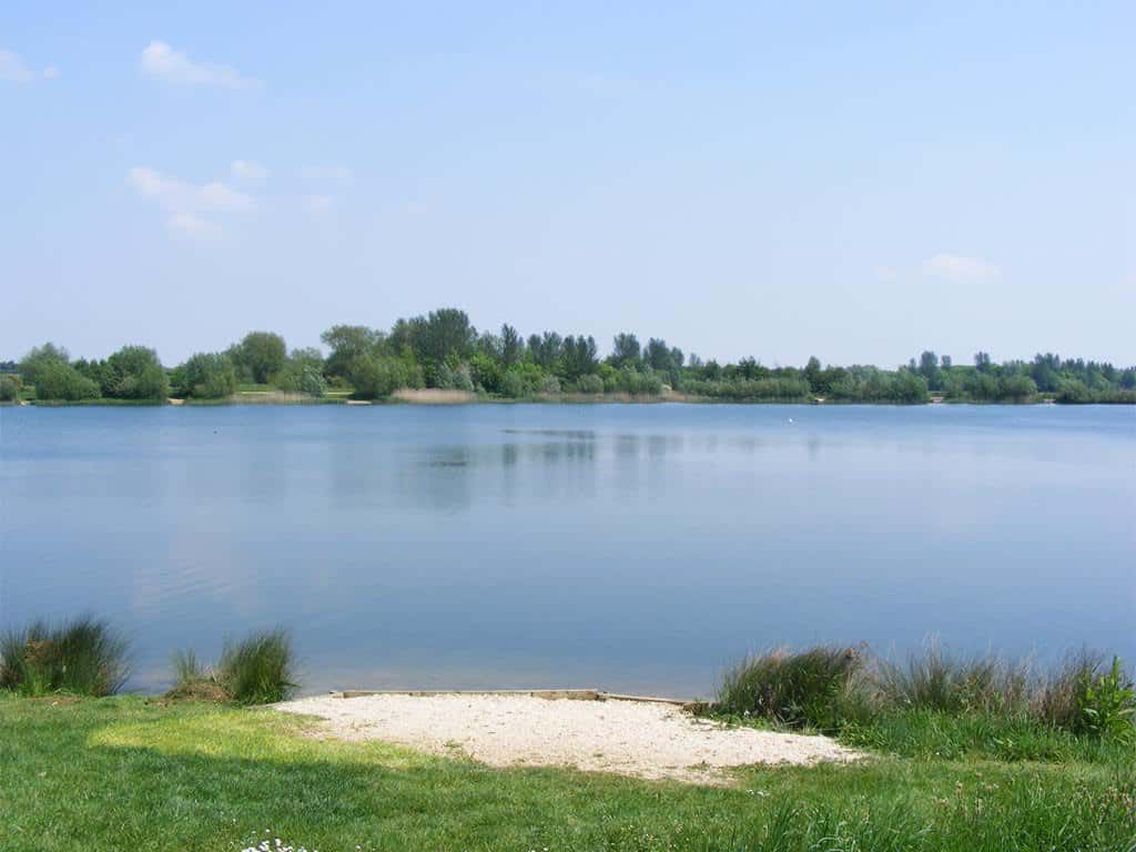 A calm lake with clear water, grassy banks, and a small sandy area in the foreground; trees and blue sky are visible in the distance.